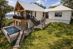 Rear exterior view of a Hill Country home with a pool deck, covered balcony, stone walls, black railings, and lake views.