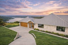 Hill Country lake-view home exterior with stone siding, a metal roof, wood entry accents, and a sunset sky.